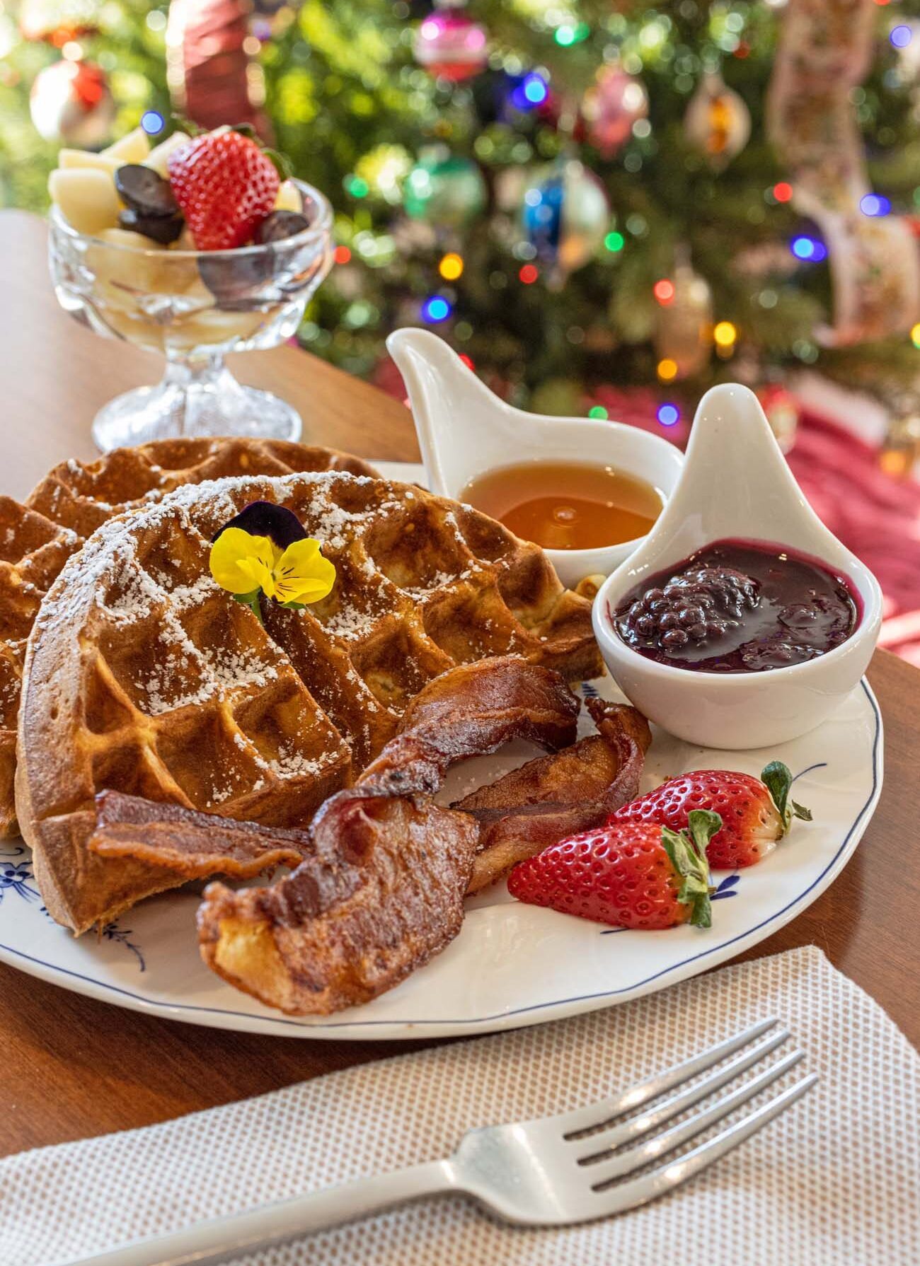 Belgian waffle dusted with powdered sugar, garnished with yellow pansy flower, served with crispy bacon strips, fresh strawberry, fruit compote, maple syrup, and jam in white ceramic dishes, Christmas tree with colored lights blurred in background