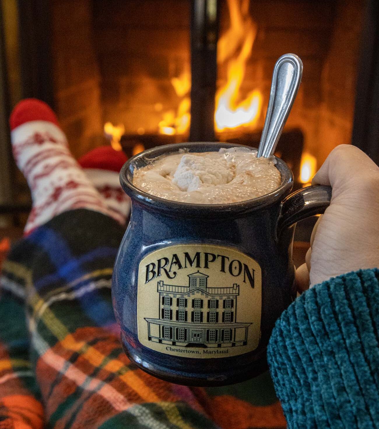 Brampton-branded blue ceramic mug filled with hot chocolate topped with whipped marshmallow cream in foreground, person in plaid flannel blanket relaxing by roaring fireplace with orange and yellow flames