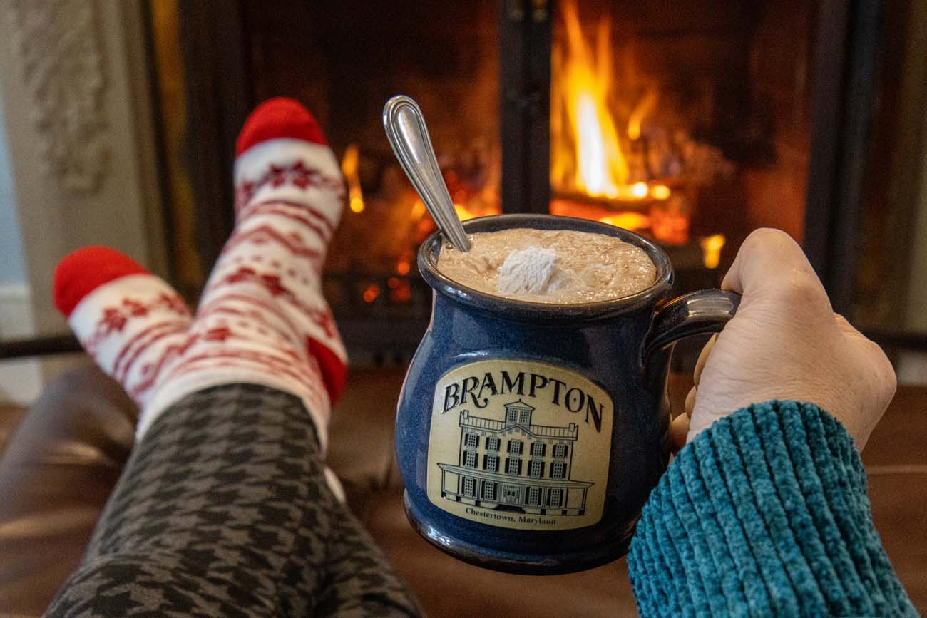 Person in Fair Isle sweater and red snowflake socks relaxing by fireplace holding Brampton-branded blue ceramic mug of hot chocolate topped with marshmallow, cozy winter scene with plaid blanket and warm fire