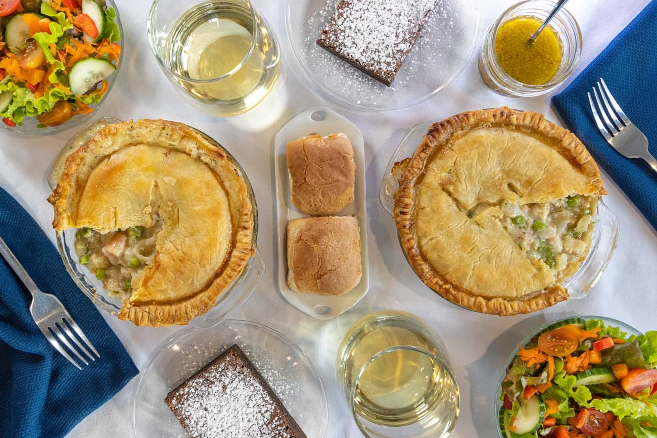 Two individual seafood pot pies with golden flaky pastry crusts in ceramic dishes, served with mixed green salads, white wine in stemless glasses, crusty rolls, powdered sugar-dusted brownies, overhead table setting