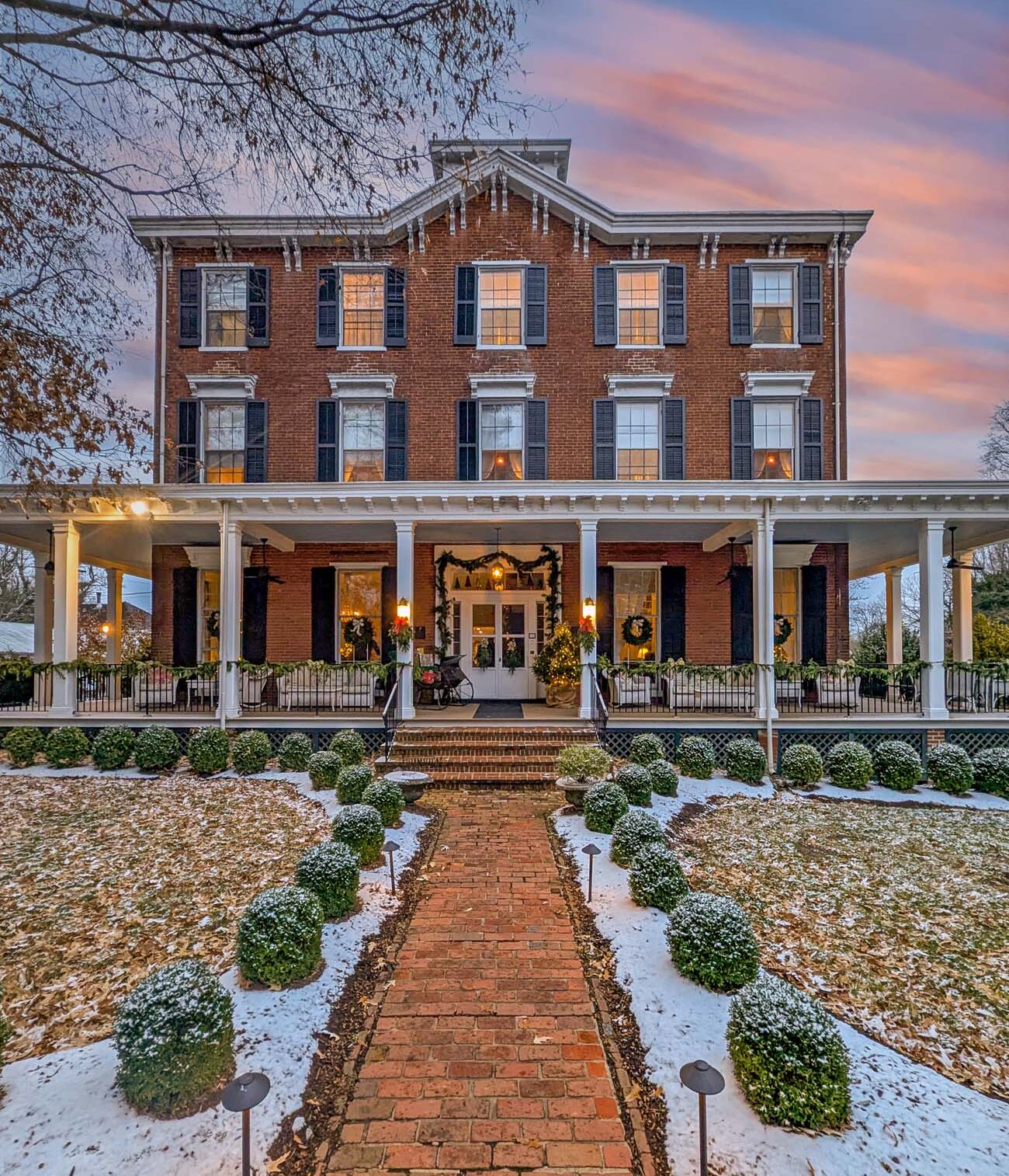 Brampton 1860 three-story brick Georgian manor house decorated for Christmas with evergreen wreaths, garland, and white lights on columned porch, dusted with fresh snow on lawn and brick walkway, warm window lights glowing against pink and purple sunset sky