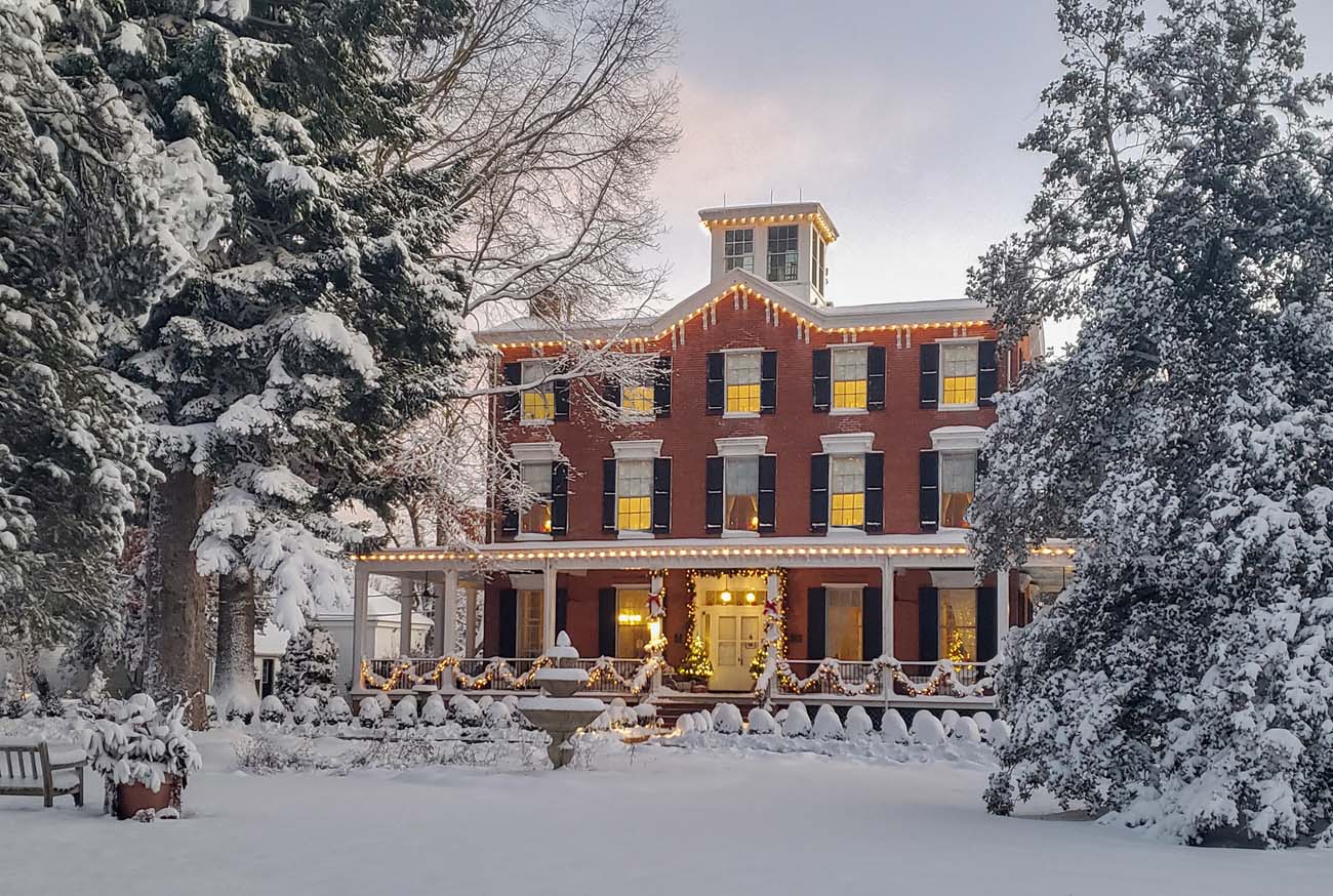 Brampton 1860 Manor House completely blanketed in heavy snow, three-story brick building with holiday lights outlining porch and roofline, evergreen wreaths on windows, snow-covered evergreen trees flanking property, warm yellow light glowing from windows in evening