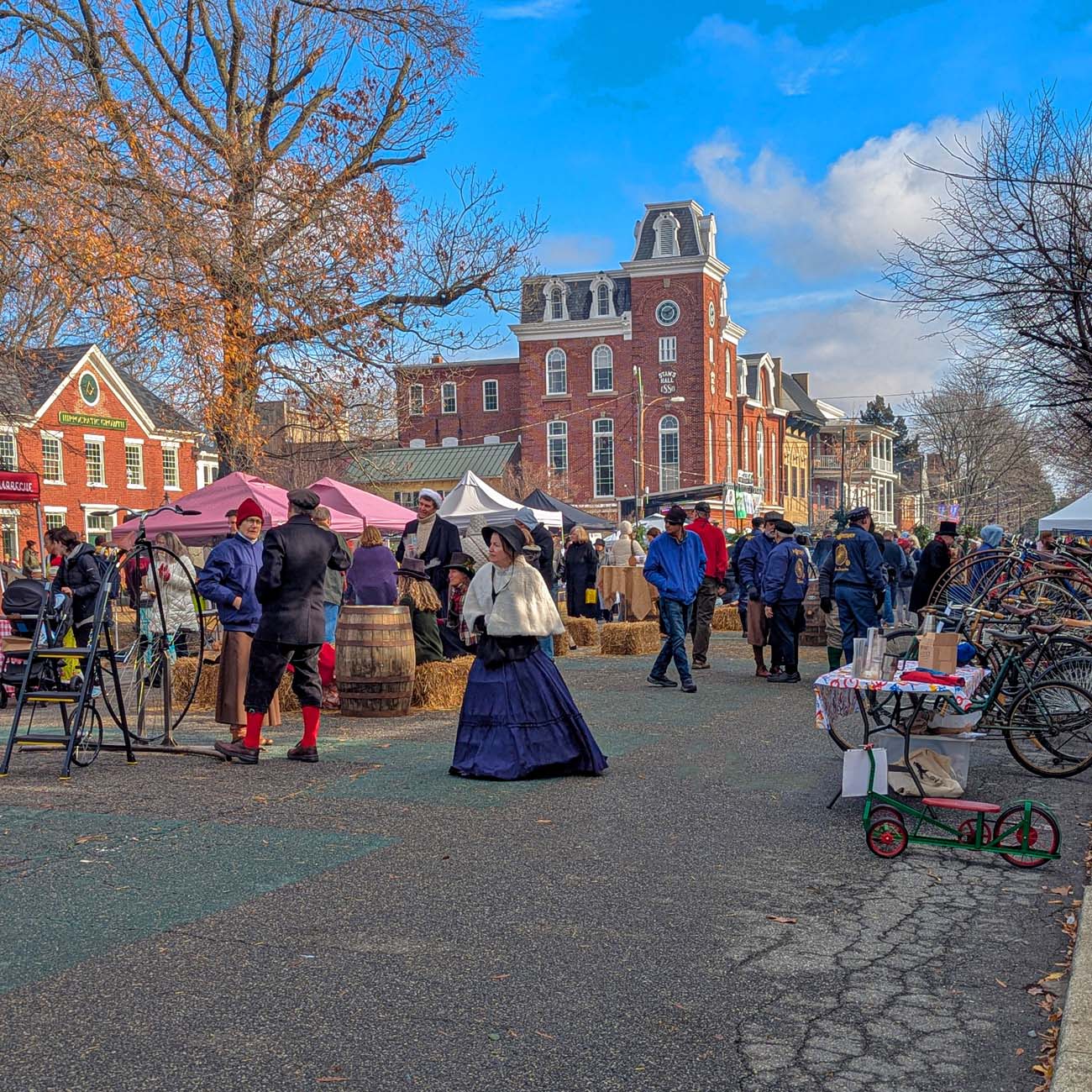 Wide view of Dickens of a Christmas festival on closed High Street showing Victorian-dressed attendees, vendor tents, hay bales, historic clock tower building in background