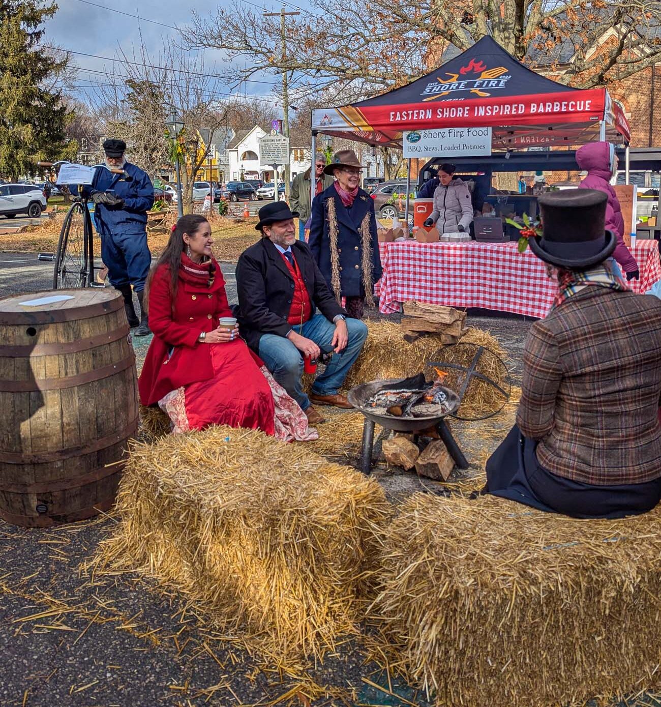 Victorian-dressed couple in period costumes sitting on hay bales around small firepit at Dickens of a Christmas festival, woman in red velvet coat with top hat, man in black suit, Eastern Shore Fire BBQ tent and food vendors in background, authentic 1840s London atmosphere