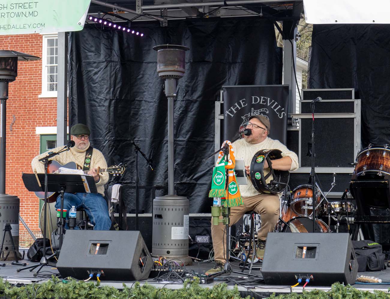 The Devil's Brigade performing on outdoor stage at Dickens of a Christmas, two musicians in cream sweaters playing guitar and bodhran drum, black backdrop with band logo, professional sound equipment, live music entertainment