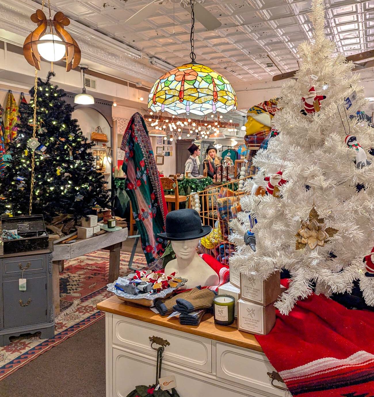 Interior of Walnut & Wool boutique in Chestertown decorated for holidays, white vintage Christmas tree with ornaments, traditional green tree with lights, Tiffany-style pendant lamp, mannequin with bowler hat displaying scarves and accessories, vintage furniture, pressed tin ceiling