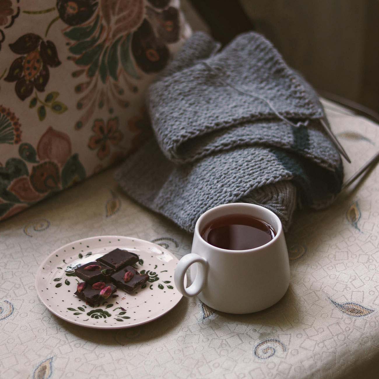 Gray knitted fabric with metal knitting needles resting on top, placed on a patterned tablecloth next to a white ceramic mug filled with tea and a small plate with chocolate pieces and dried flower petals, creating a cozy wellness scene