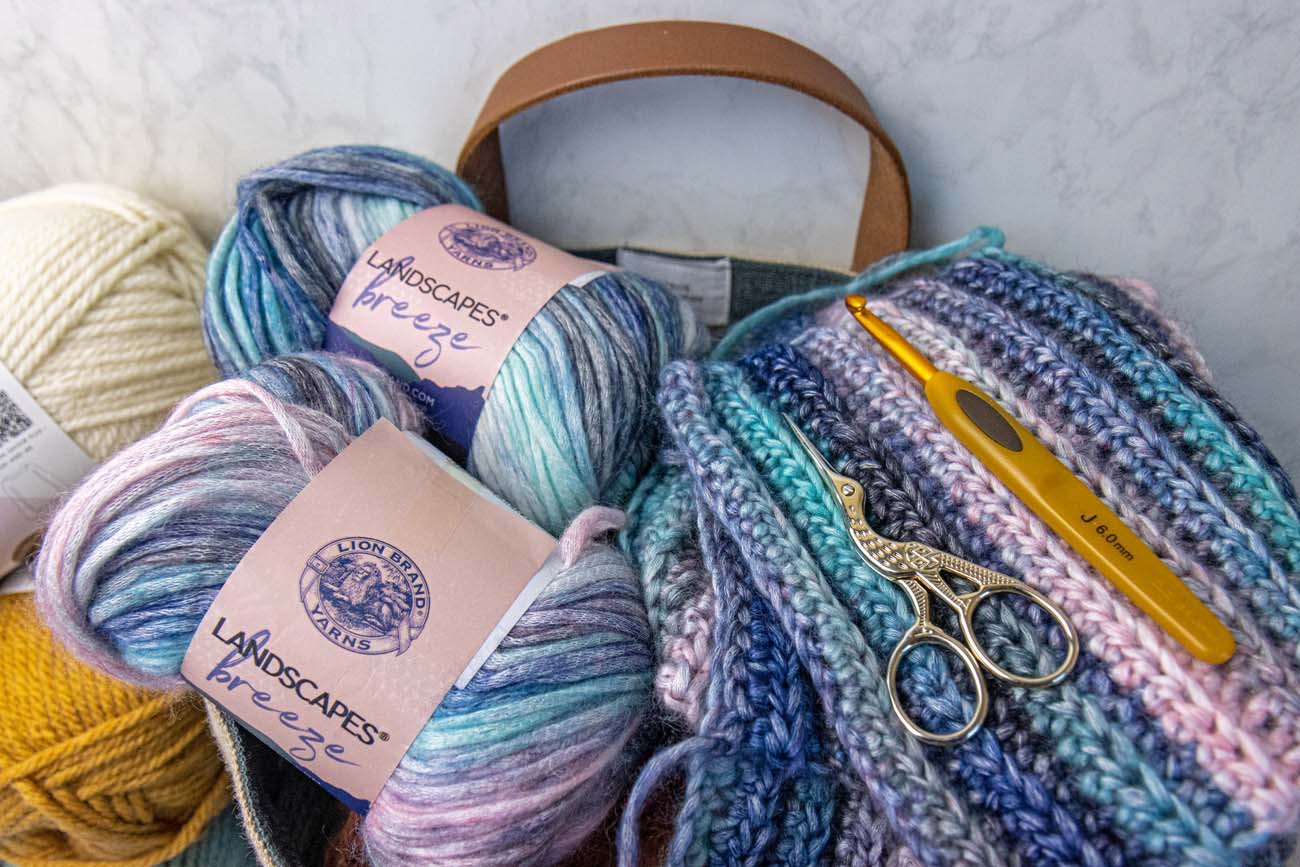 Close-up of Lion Brand Landscapes Breeze yarn skeins in coastal blue, pink, and cream tones inside a canvas tote bag with leather handles, featuring completed crochet squares and golden scissors