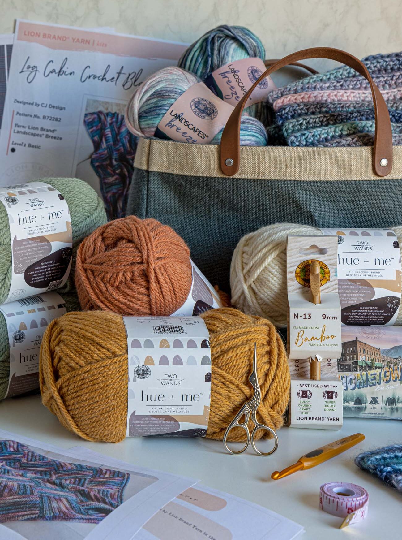 Flat lay of Lion Brand crochet kit contents including Log Cabin pattern card, canvas tote bag filled with Landscapes Breeze yarn, multiple skeins of Hue + Me yarn in orange, yellow, and green tones, gold scissors, measuring tape, bamboo crochet hooks, and completed crochet squares