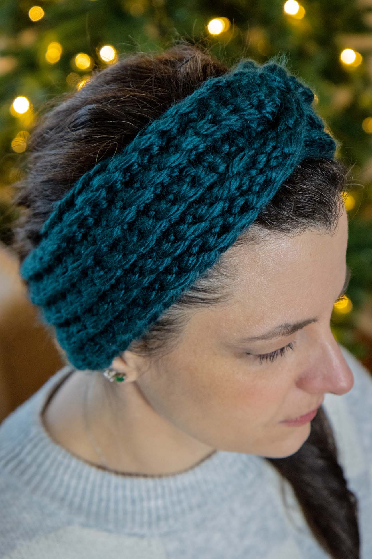 Woman wearing teal knitted headband outdoors with holiday lights bokeh in background, eyes closed in peaceful expression, wearing gray sweater