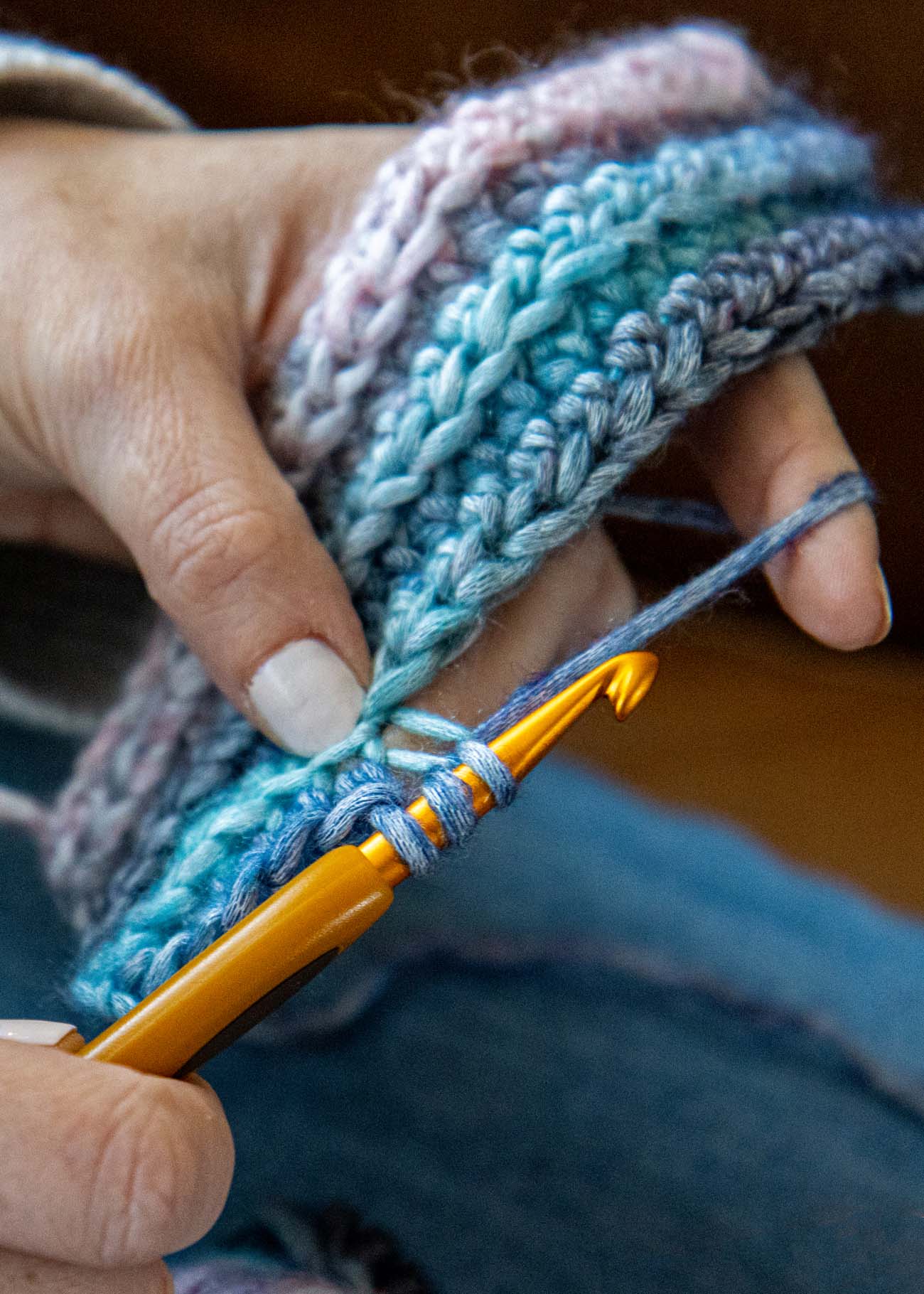 Hands holding a crochet work in progress with gold crochet hook, showing coastal-toned stitches in blue, pink, and cream from Lion Brand Landscapes Breeze yarn, skein visible in foreground