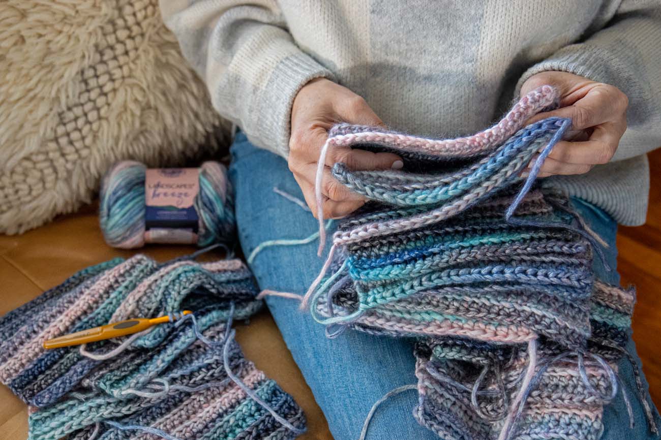 Overhead view of hands holding stack of crocheted squares in coastal blues, pinks, and grays on lap with Lion Brand Landscapes Breeze yarn and gold crochet hook visible, sheepskin rug in background