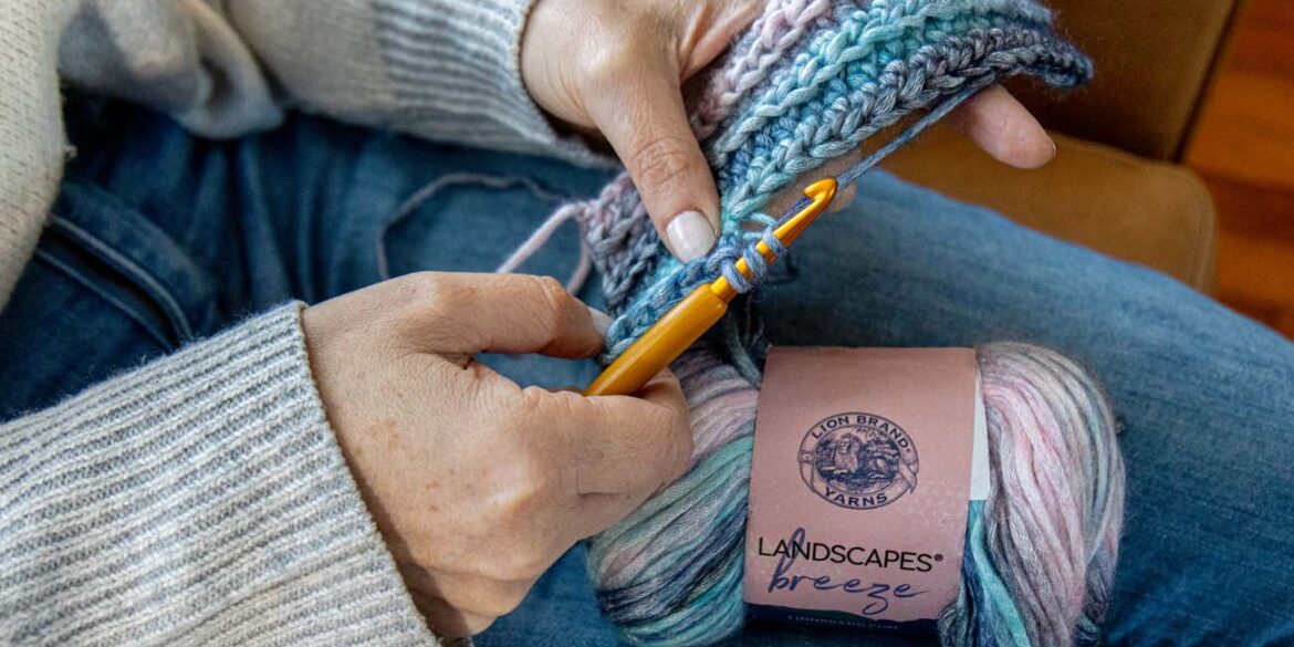 Hands crocheting with gold hook and Lion Brand Landscapes Breeze yarn in coastal tones, showing active stitching technique with yarn skein resting on denim-clad lap