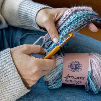 Hands crocheting with gold hook and Lion Brand Landscapes Breeze yarn in coastal tones, showing active stitching technique with yarn skein resting on denim-clad lap
