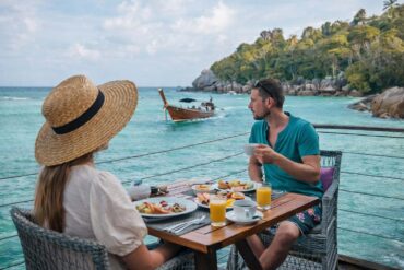 a man on woman dining dockside in Thailand as a wooden boat drifts by
