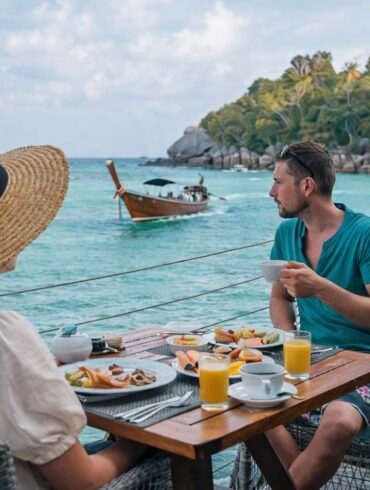 a man on woman dining dockside in Thailand as a wooden boat drifts by