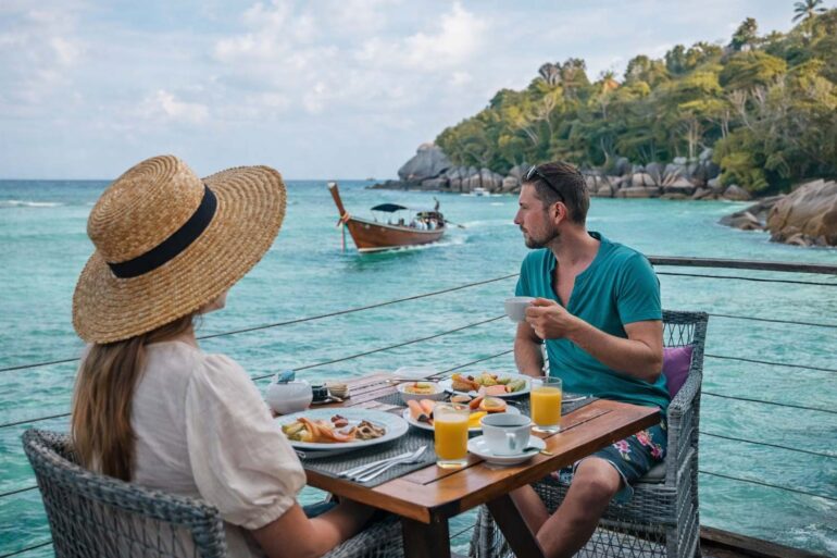 a man on woman dining dockside in Thailand as a wooden boat drifts by
