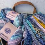 Lion Brand Landscapes Breeze gradient yarn skeins in coastal blues, pinks, and creams with completed crochet squares, gold scissors, and yellow crochet hook in canvas tote bag for our Lion Brand Yarn Review