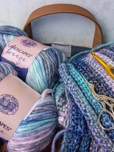 Lion Brand Landscapes Breeze gradient yarn skeins in coastal blues, pinks, and creams with completed crochet squares, gold scissors, and yellow crochet hook in canvas tote bag for our Lion Brand Yarn Review