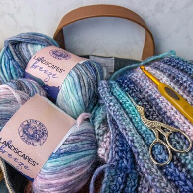 Lion Brand Landscapes Breeze gradient yarn skeins in coastal blues, pinks, and creams with completed crochet squares, gold scissors, and yellow crochet hook in canvas tote bag for our Lion Brand Yarn Review