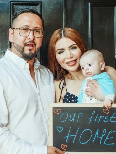 a man, woman, and infant in front of their new home with a sign that says our first home