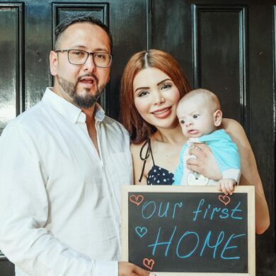 a man, woman, and infant in front of their new home with a sign that says our first home