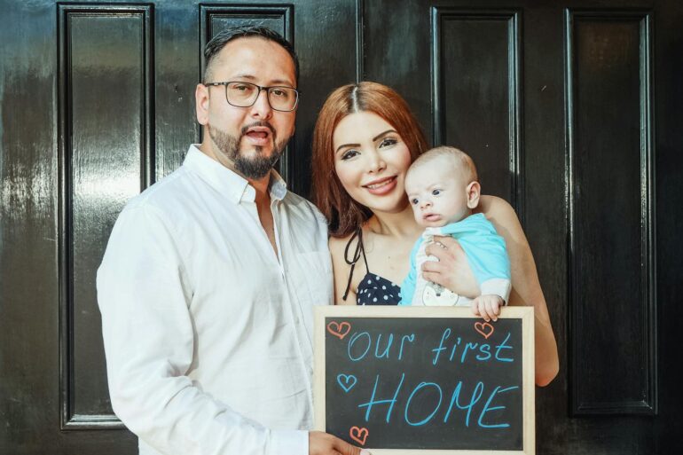 a man, woman, and infant in front of their new home with a sign that says our first home