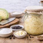 Glass fermentation jar filled with homemade sauerkraut showing golden fermented cabbage submerged in cloudy brine, surrounded by fresh green cabbage, sea salt in white bowl, juniper berries, caraway seeds in wooden spoon, peeled garlic cloves, and peppercorns on rustic wooden cutting board with burlap cloth and ceramic crocks in background