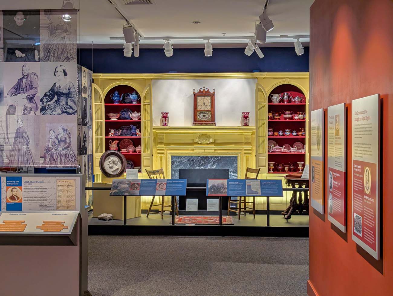 An exhibit room inside the Chester County History Center featuring a reconstructed yellow Federal-style mantelpiece with a marble fireplace surround and arched built-in shelving filled with red and blue historic ceramics, silver pieces, and an antique clock. Blue interpretive panels stand in the foreground, and the left wall displays large-format historic photographs and a biographical panel about Uriah Hunt Painter. Additional exhibit panels about civil rights history line the red-painted right wall.