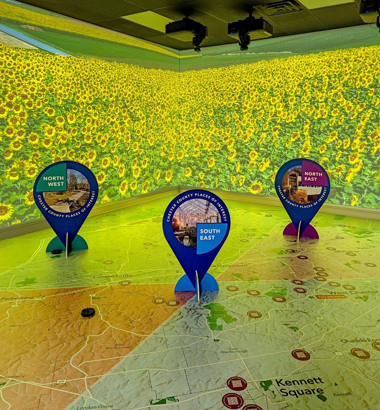 The immersive map room inside the Chester County Welcome Center, with a floor-to-ceiling projection of a sunflower field on the surrounding walls and a large printed map of Chester County on the floor. Three life-sized blue location-pin markers labeled North West, South East, and North East stand on the map, each featuring a circular photo of a local attraction.