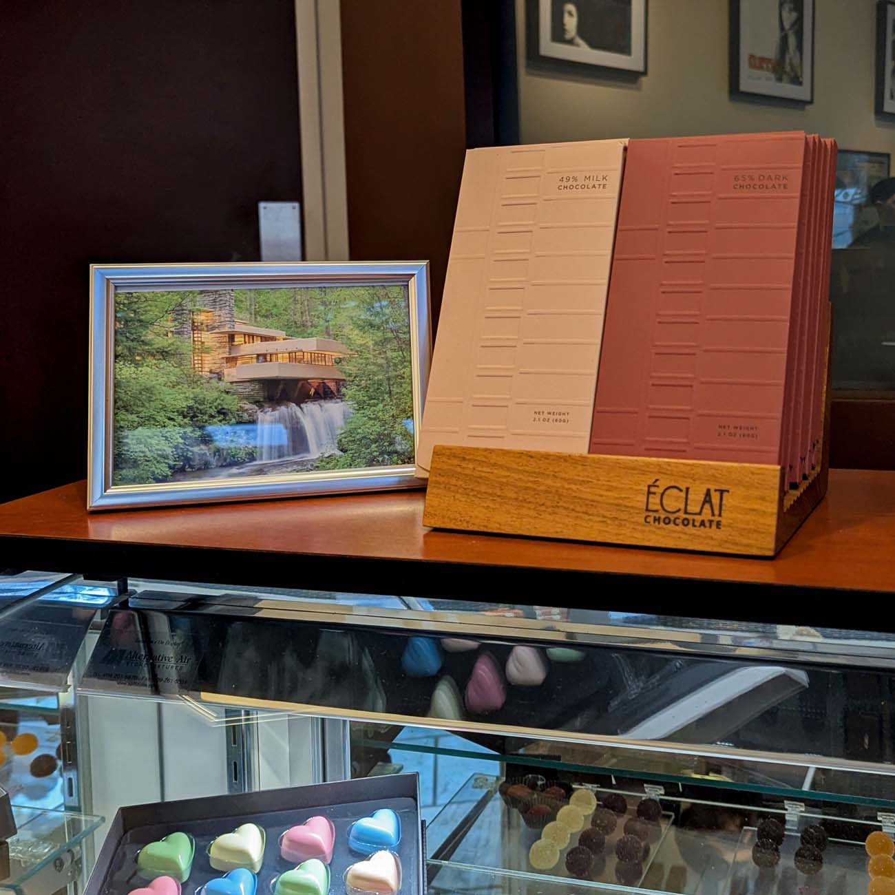 A display inside Eclat Chocolate in West Chester showing the signature Fallingwater Bars in milk chocolate and dark chocolate varieties standing in a wooden holder branded with the Eclat Chocolate logo. A framed photograph of Frank Lloyd Wright's Fallingwater house sits beside the bars. Below the glass counter, rows of colorful heart-shaped bonbons and hand-painted chocolates are visible.