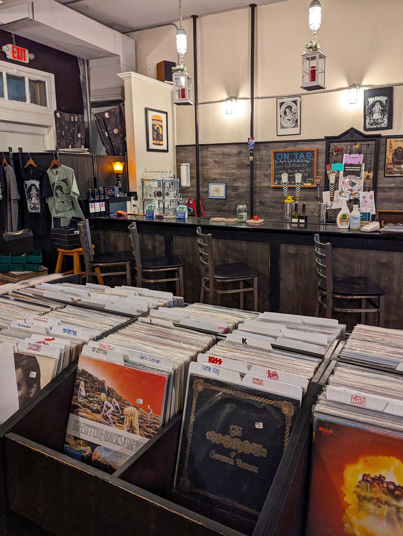 Bins of vinyl records in the foreground at Hop Fidelity in West Chester, with visible album covers including Led Zeppelin, Kiss, and other classic rock titles. The nano brewery bar with stools, an "On Tap" chalkboard, hanging lantern-style lights, and framed music posters line the back wall. Band t-shirts hang on a rack near the entrance.