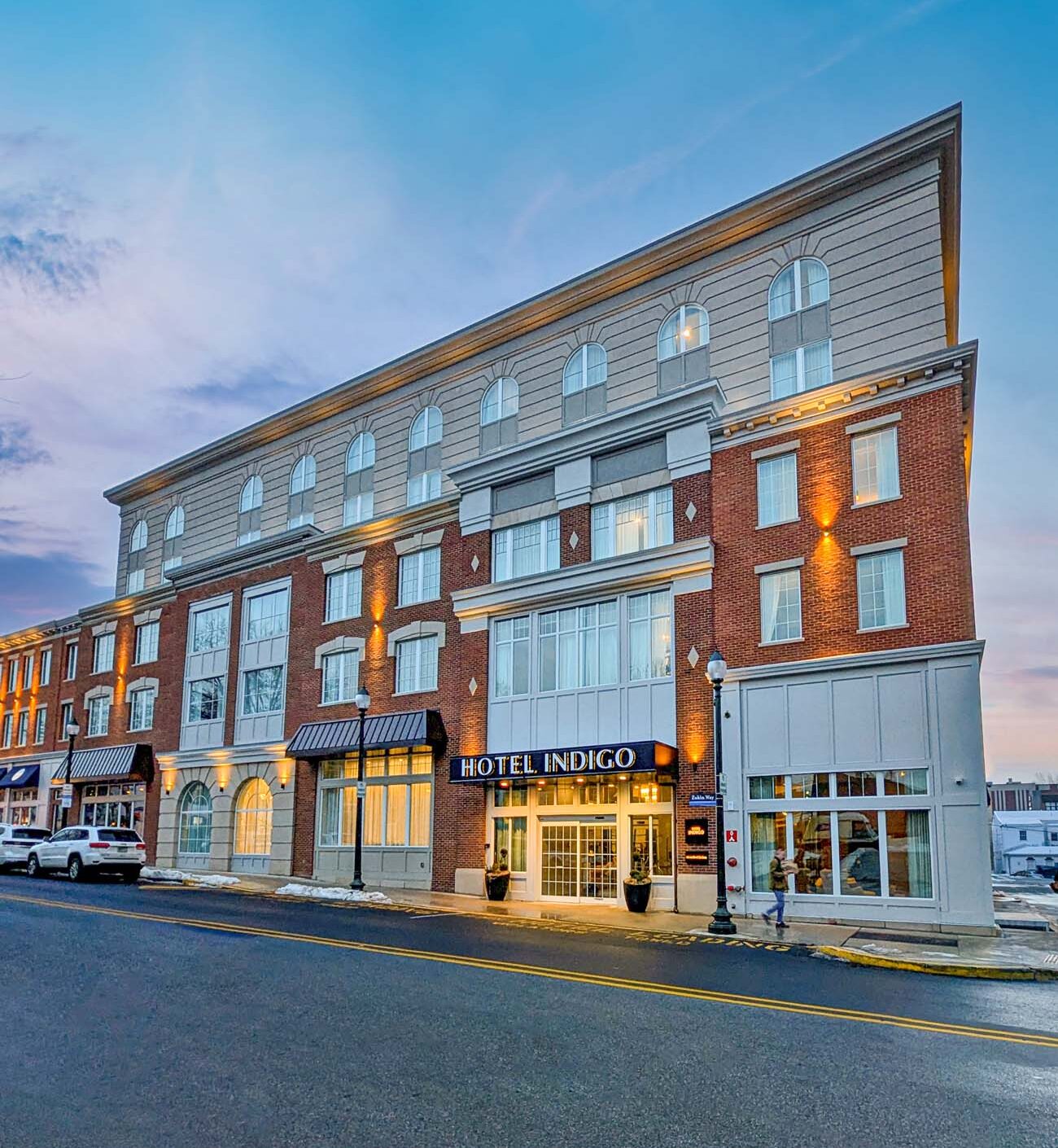 The exterior of Hotel Indigo West Chester at dusk, a four-story red brick and gray facade building with arched upper-floor windows, warm exterior lighting, and a dark awning over the main entrance reading "Hotel Indigo." The street is quiet with a few parked cars and a pastel blue and pink sky above.