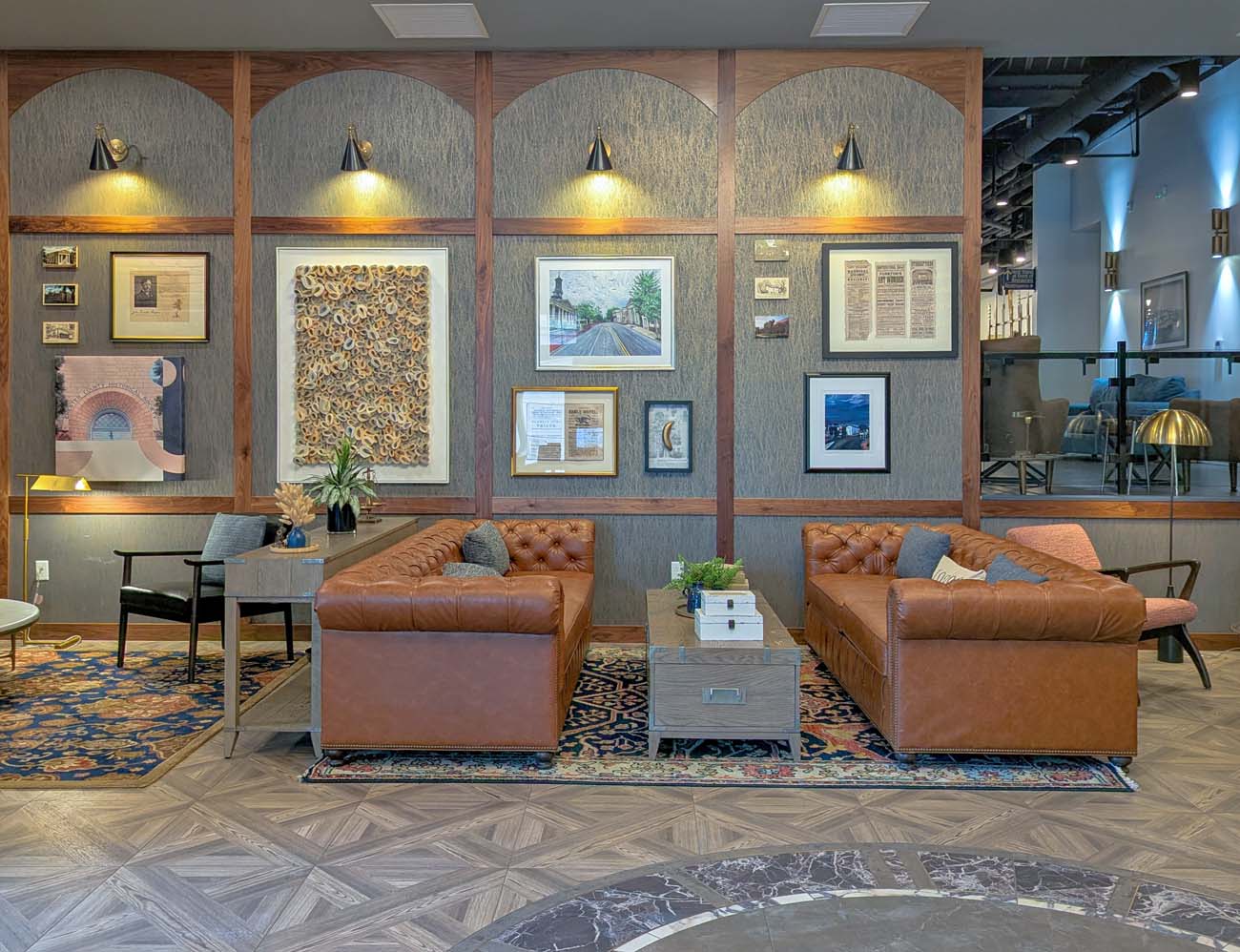 A seating area in the Hotel Indigo West Chester lobby with two tufted brown leather sofas facing each other across a gray wooden coffee table, set on a herringbone wood floor with a circular marble inlay. The wall behind features arched wood-framed panels with a gallery of local artwork, historic prints, and photographs, lit by black and brass cone sconces. A raised lounge area is visible in the background.