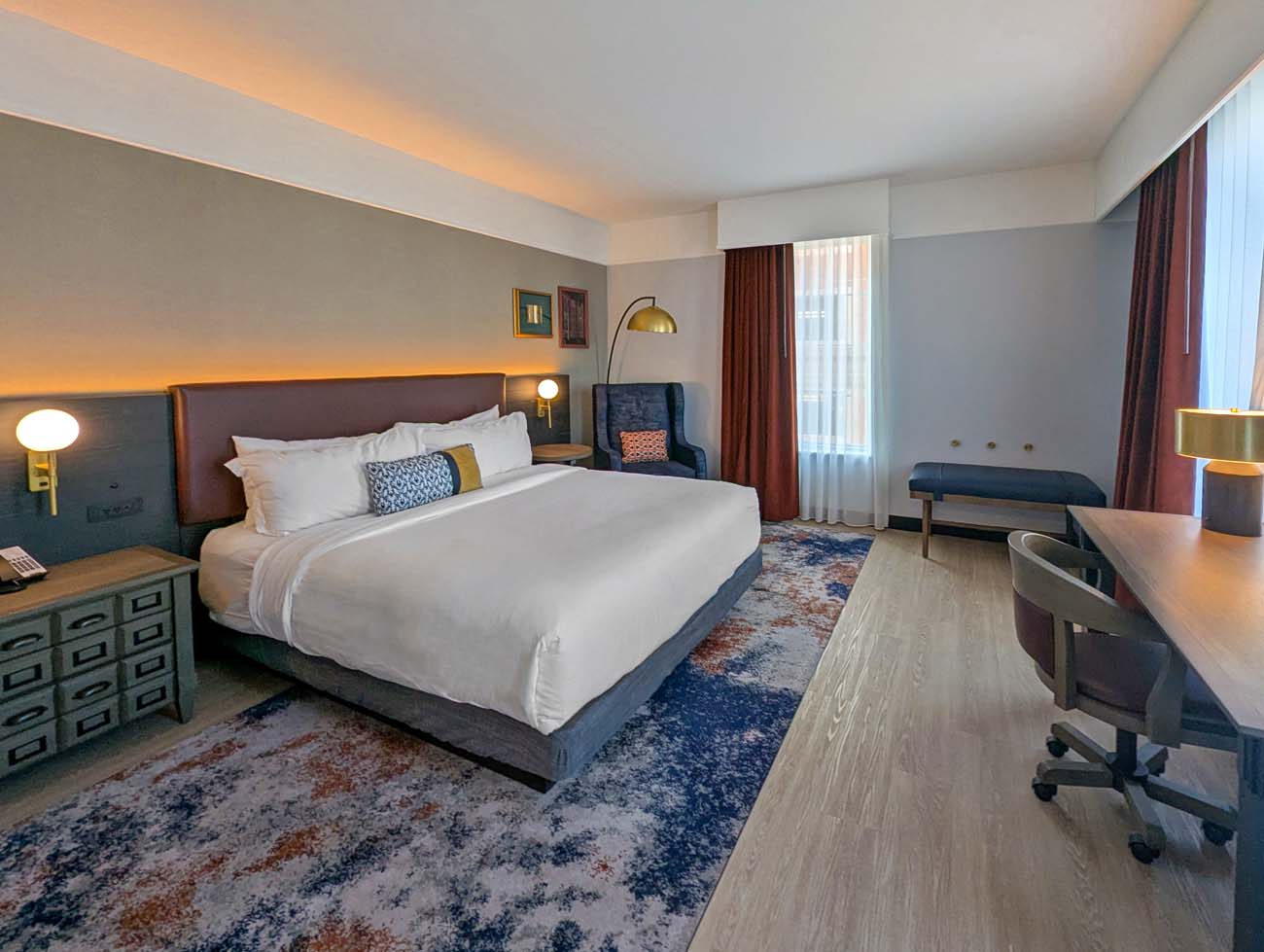 A spacious corner king suite at Hotel Indigo West Chester with a large bed dressed in white linens and blue accent pillows, warm overhead cove lighting, a navy wingback chair beside the bed, a long wooden desk with a rolling chair, a navy bench along the window wall, and a blue and copper abstract area rug on light hardwood floors. Rust-colored blackout curtains frame the window.