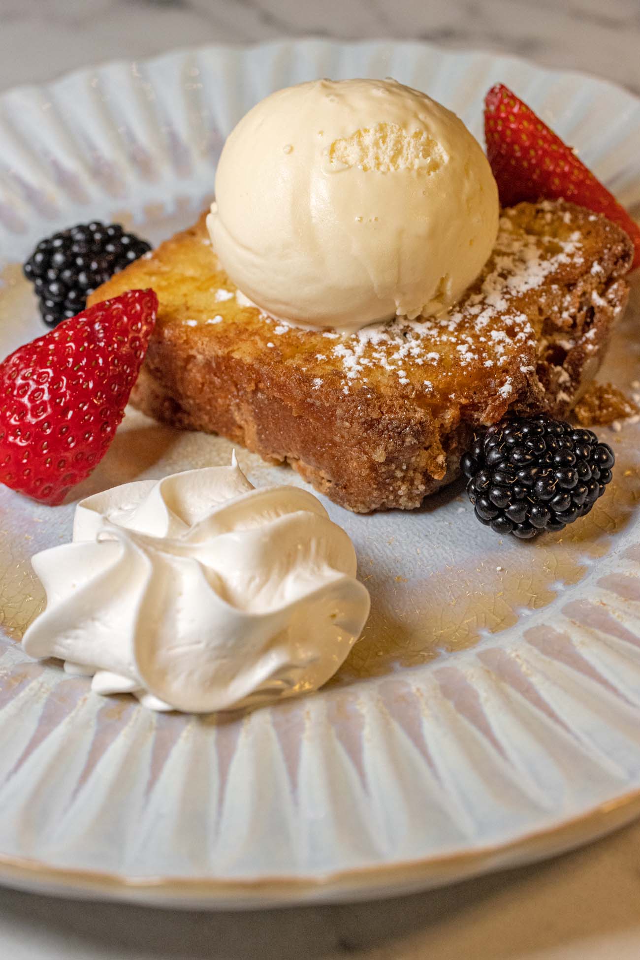 A slice of Limoncello Cake on a fluted white and gold plate, topped with a scoop of vanilla gelato and dusted with powdered sugar. Fresh strawberry halves and whole blackberries surround the cake, with a rosette of whipped cream piped to one side.