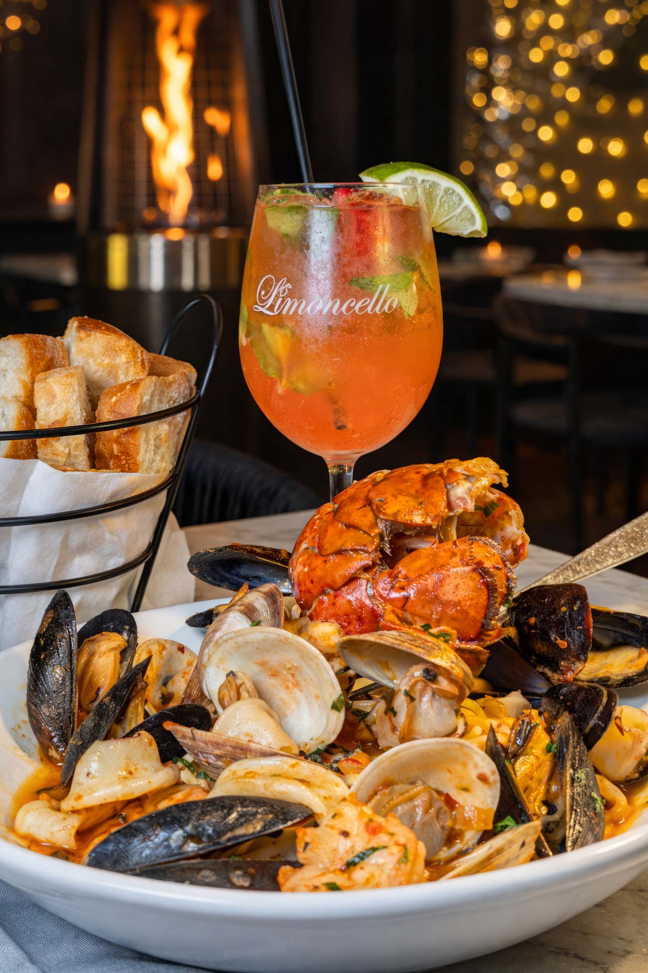 A large white bowl of Pescatore Fra Diavolo at Limoncello Ristorante in West Chester, piled high with a split lobster tail, mussels, clams, shrimp, and calamari over linguine in a red spicy tomato broth. A branded Limoncello cocktail glass filled with an orange spritz and garnished with a lime wedge sits behind the dish, with a wire basket of sliced bread and a flame heater glowing with fairy lights in the background.