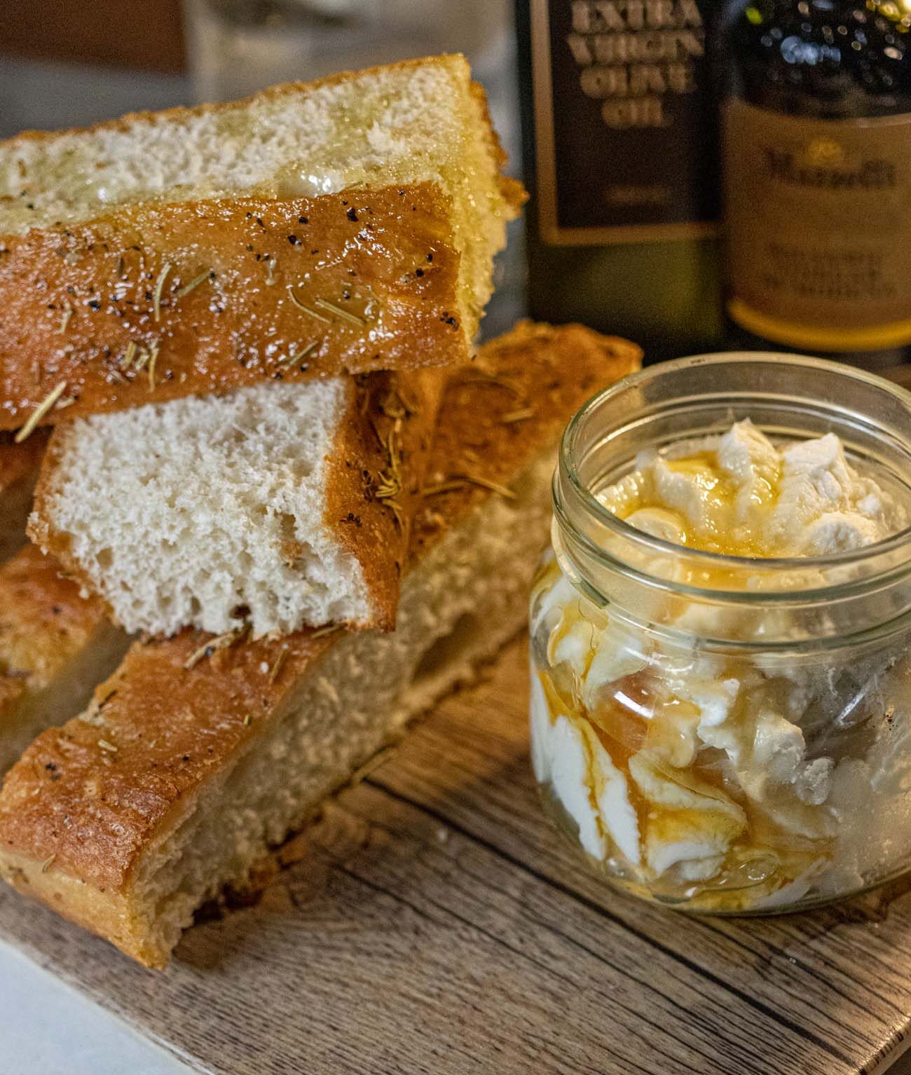 The Ricotta Board at Limoncello Ristorante, served on a wooden plank with thick slices of golden rosemary focaccia stacked beside a small mason jar of fresh ricotta drizzled with honey. A bottle of extra virgin olive oil and balsamic vinegar sit in the background on a marble tabletop.