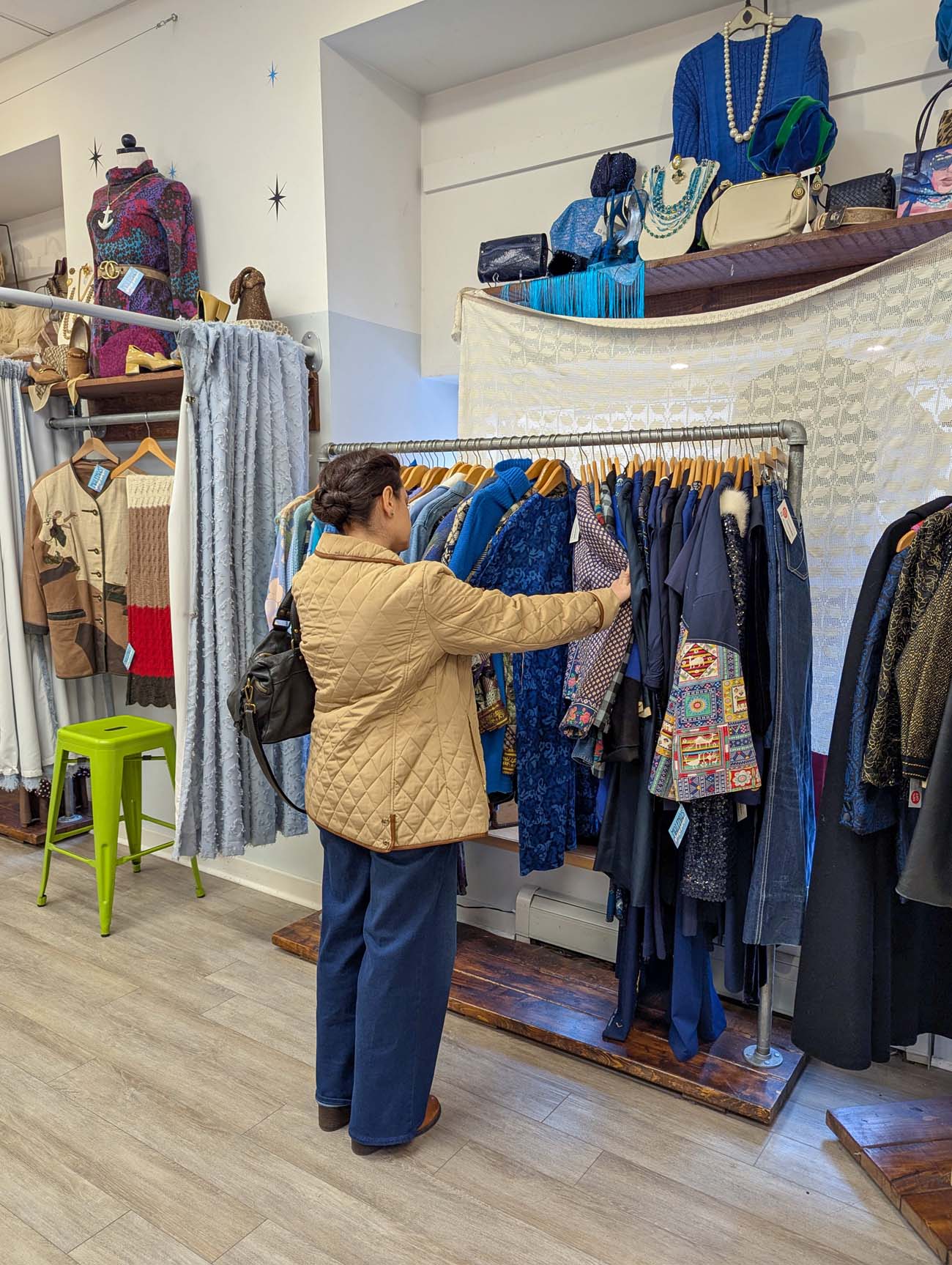 A shopper browsing a rack of vintage clothing organized by color at Malenas Vintage Boutique in West Chester. The rack features blues, navy, and patterned pieces from various eras. Shelves above display vintage accessories, jewelry, and handbags. A green metal stool and a gray curtained fitting area are visible to the left, and more colorful vintage garments and mannequins fill the background.