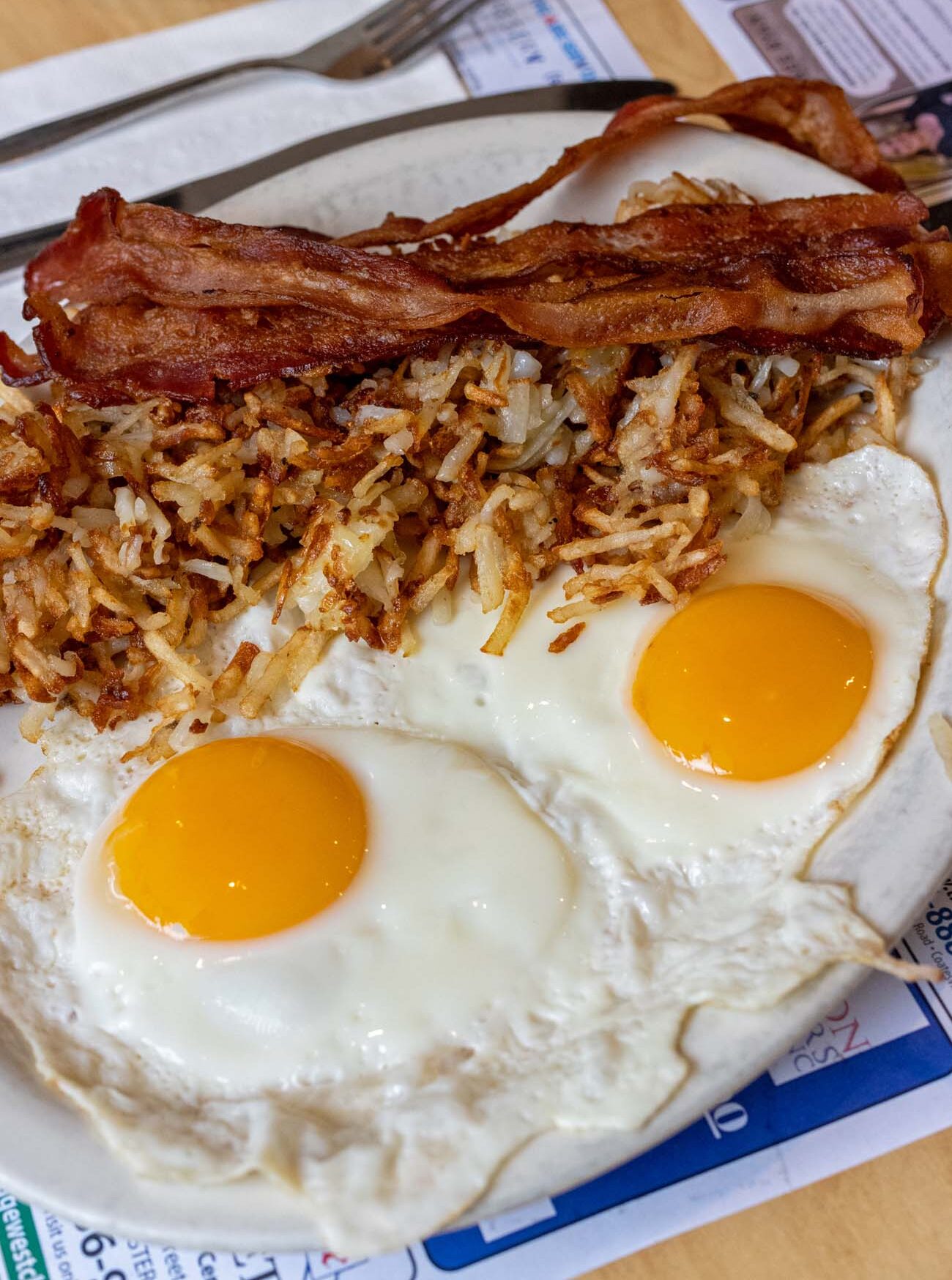 A breakfast plate at Market Street Grill with two sunny-side-up eggs with bright orange yolks, a pile of golden crispy hashbrowns, and two strips of thick-cut bacon, served on a white plate atop a paper placemat printed with local advertisements.