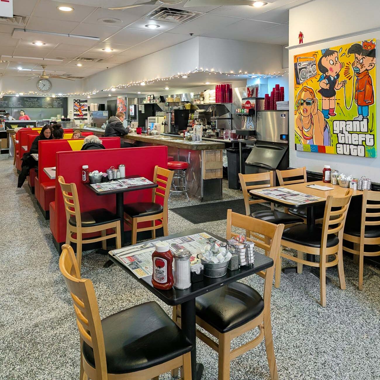 The interior of Market Street Grill in West Chester, a classic diner with red vinyl booths along the left wall, wooden tables and chairs in the center, a long counter with stools, string lights above the kitchen area, and colorful original artwork hanging on the walls including a large pop-art style painting. A few morning diners are seated at the booths.