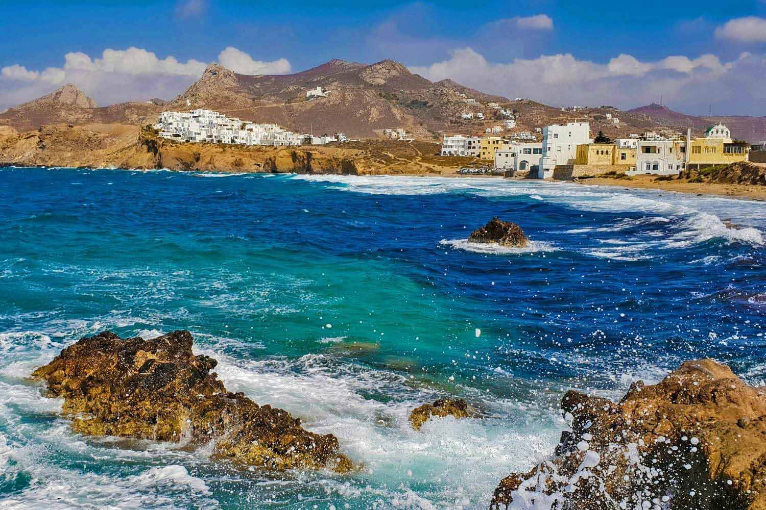 A view of Naxos Greece from the water. Mountains and quaint villages in the distance as the tide breaks on the rock formations a few yards out from shore.