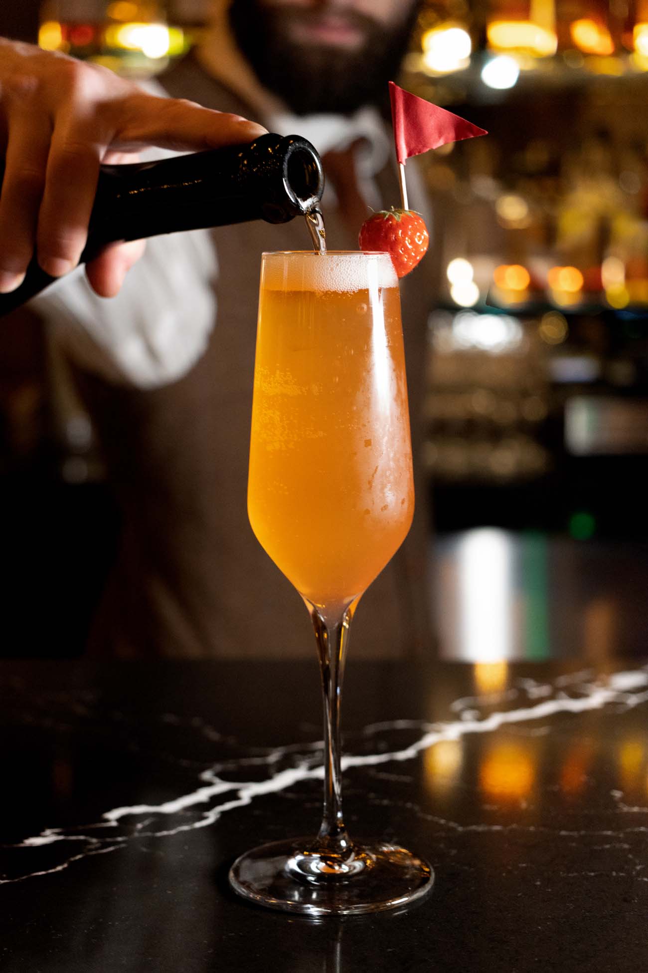 A bartender pouring prosecco from a dark bottle into a tall flute glass filled with an orange cocktail garnished with a fresh strawberry and a small red paper flag on a cocktail pick. The glass sits on a black marble bar top with warm, softly blurred bar lighting in the background.
