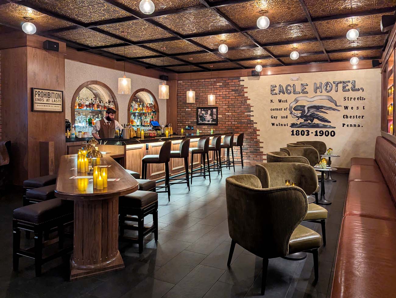  The interior of Room 109 Speakeasy beneath Hotel Indigo in West Chester, featuring a full bar with arched backlit shelving stocked with spirits, leather and wood bar stools, olive green velvet lounge chairs at small candlelit tables, a long leather banquette along the right wall, and a large painted mural of the historic Eagle Hotel reading "Eagle Hotel, N. West corner of Gay and Walnut Streets, West Chester Penna. 1803–1900." An embossed tin ceiling with globe lights runs overhead, and a framed "Prohibition Ends at Last!" sign hangs on the left wall. A bartender in a vest stands behind the bar.
