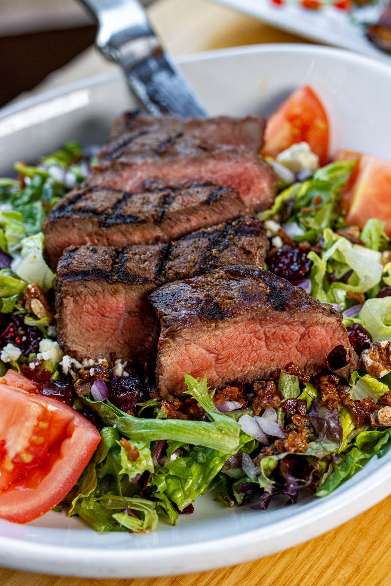 A Gorgonzola Chopped Salad with Steak at Sedona Taphouse, featuring thick slices of grilled medium-rare steak with dark char marks laid over a bed of chopped mixed greens, crumbled gorgonzola, diced red onion, dried cranberries, bacon bits, and tomato wedges in a white bowl.