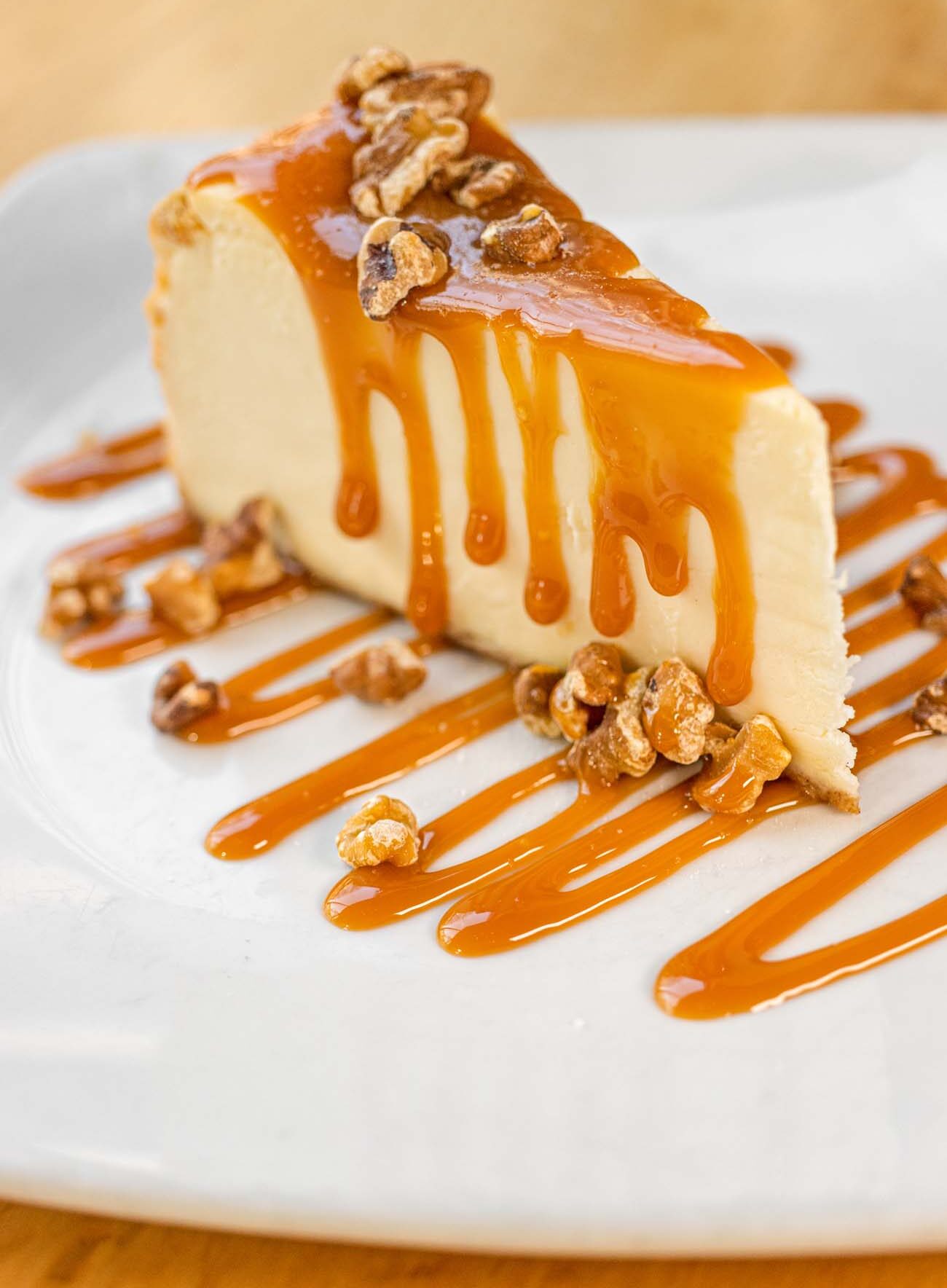A generous slice of Salted Caramel Cheesecake at Sedona Taphouse, smooth and pale with a thick layer of caramel sauce dripping down the sides, topped and surrounded by candied nuts, on a white square plate with caramel drizzled in parallel lines beneath the slice.
