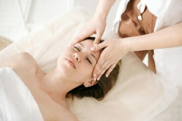 A licensed skincare professional performing a facial treatment on a relaxed woman lying on a treatment table at a medical aesthetic clinic, with gentle hand placement on her forehead in a clean, softly lit spa environment.