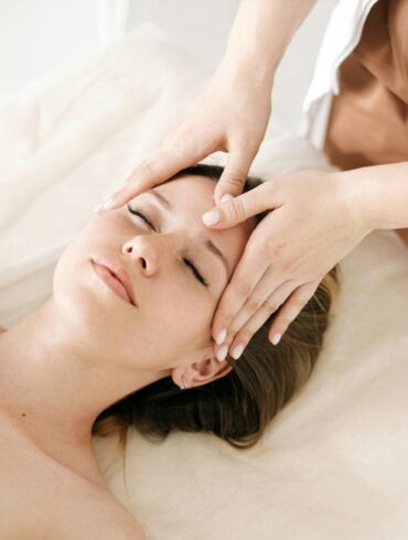 A licensed skincare professional performing a facial treatment on a relaxed woman lying on a treatment table at a medical aesthetic clinic, with gentle hand placement on her forehead in a clean, softly lit spa environment.