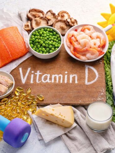 Overhead photo of vitamin D foods and fitness items arranged on a light background. In the center is a wooden board with the words ‘Vitamin D’ written on it. Around the board are salmon, a whole fish, shrimp, eggs, mushrooms, green peas, cheese, butter, and a glass of milk. Nearby are yellow vitamin D gel capsules and a colorful dumbbell representing exercise and muscle building. A small plush sun toy appears in the corner to represent sunlight as a source of vitamin D.