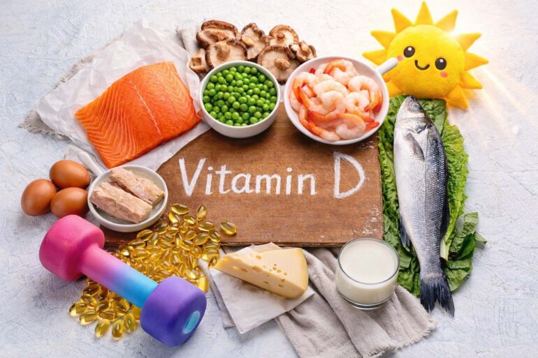 Overhead photo of vitamin D foods and fitness items arranged on a light background. In the center is a wooden board with the words ‘Vitamin D’ written on it. Around the board are salmon, a whole fish, shrimp, eggs, mushrooms, green peas, cheese, butter, and a glass of milk. Nearby are yellow vitamin D gel capsules and a colorful dumbbell representing exercise and muscle building. A small plush sun toy appears in the corner to represent sunlight as a source of vitamin D.